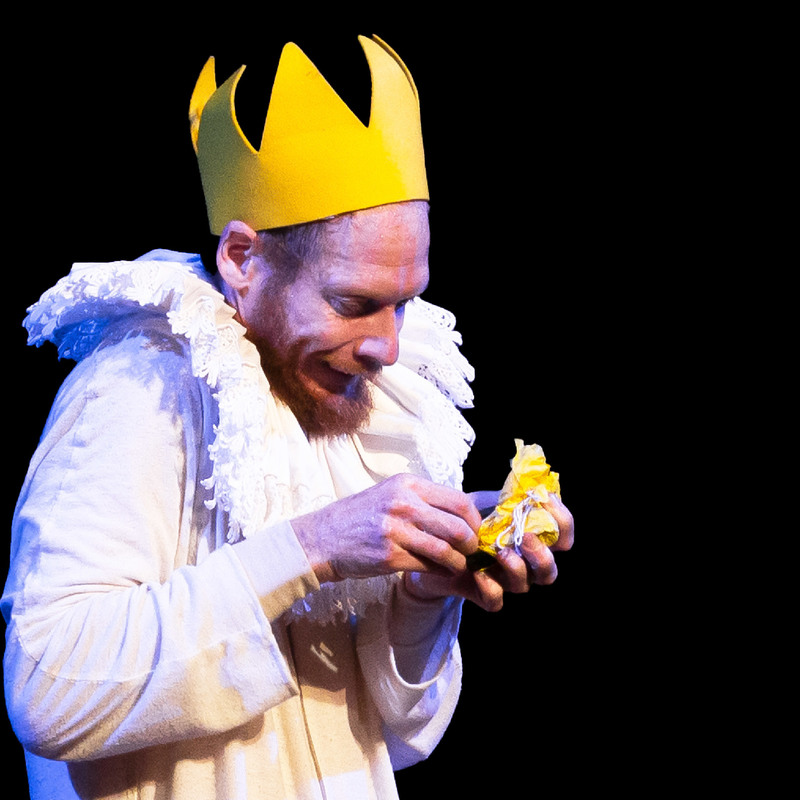Performer Thom Monckton wears a yellow felt crown and excitedly unwraps a yellow present.