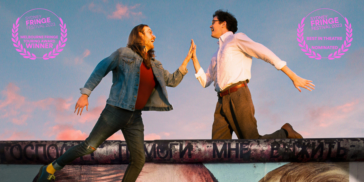 BERLINERS - Two men float towards each other above the Berlin Wall set against a peach sunset sky. The man on the left has long brown hair, and wears double denim with a red turtleneck and Doc Marten boots. The man on the right has short brown hair, and wears a simple white button-down shirt with old fashioned brown pants.