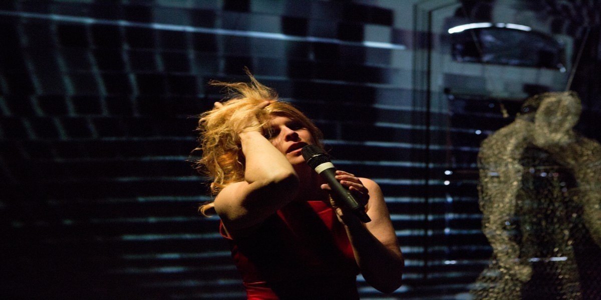 A blond woman has her eyes closed. In one hand, she has a microphone held to her mouth and the other hand is grabbing her hair. There is a silver mesh, ghostly figure next to her.