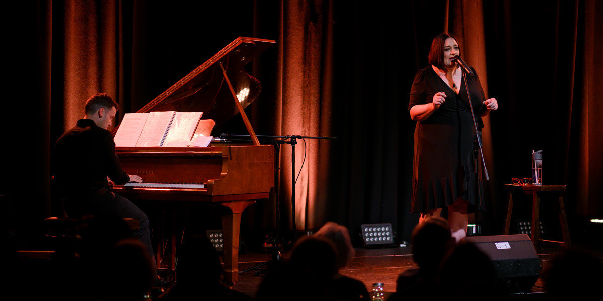Ed Heddle and Alexandra Frost - Live performance shot during Hear My Voice , Cabaret Show, with Ed engrossed on the grand piano and Alexandra, jazz, cabaret singer dressed in black and speaking into the microphone on stage at Nexus Arts, Adelaide, SA, Australia