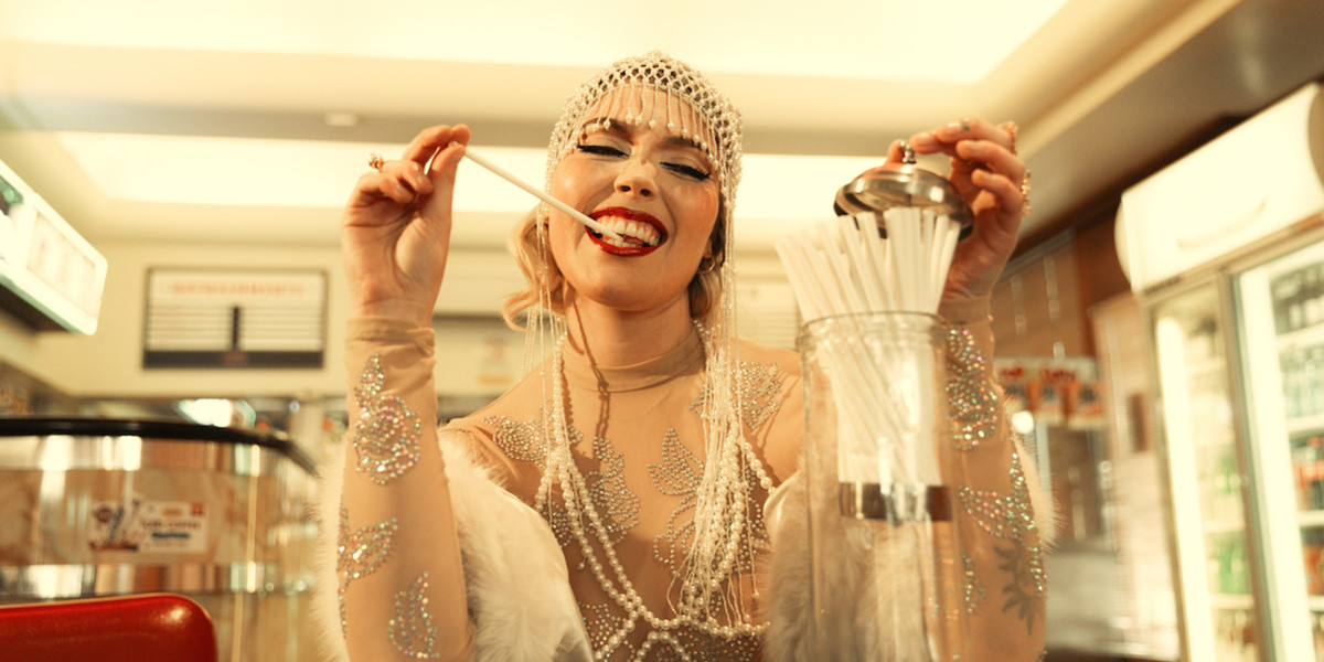 A woman wearing a white sparkling beaded headpiece and a white sparkling beaded outfit is opening a drinking straw dispenser with her left hand and has a drinking straw held between her teeth, held with her right hand. She is smiling.