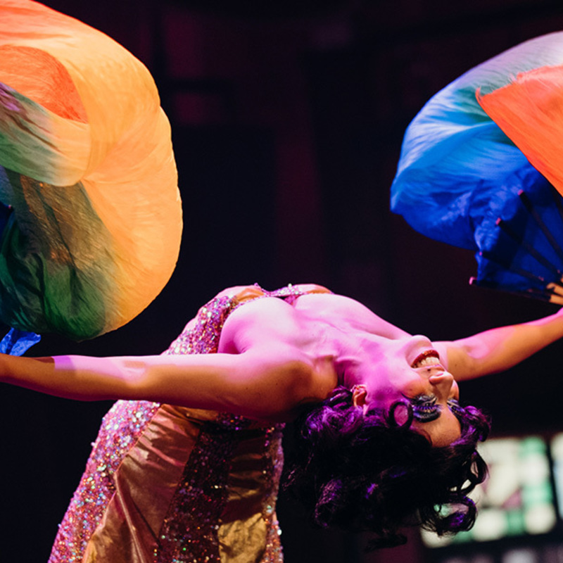 Jazida leans into a deep backbend waving 2 colourful silk fans in each hand. In the background is the stained glass of the Speigeltent.