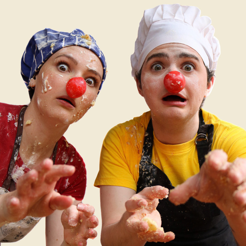 Two clowns face the camera and reach out. It is set on an off white background and it is a mid shot. The clown on the left is wearing a red short-sleeved shirt, a blue chef's hat with white stripes and a white and brown apron- she has a shocked/worried expression with mouth open. The clown on the right is wearing a yellow short-sleeved shirt, a tall white chef's hat and a black apron- they have a shocked/aghast expression with mouth open. Both are wearing red clown noses and have cake and cream smeared over their clothes. The clown on the left has cake over her hat, face and body. The clown on the right has flour over their face and cake on their hands.