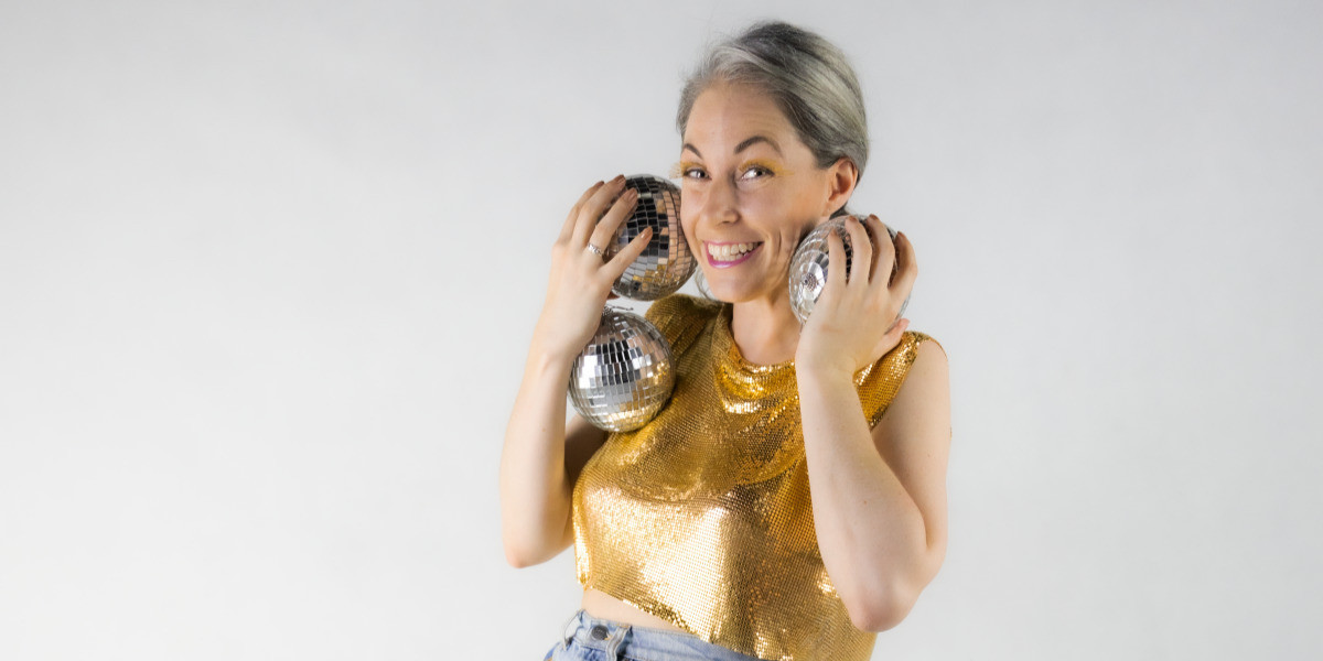 Woman smiles at camera in a gold shirt, holding mirror balls to her face