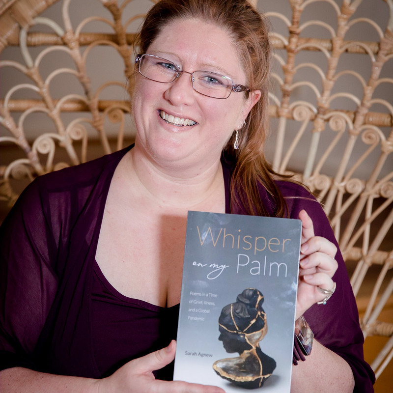 mid-40s Anglo woman wearing glasses, a burgundy blouse, and assorted jewellery, seated in a cane peacock chair, holding a copy of her book Whisper on My Palm
