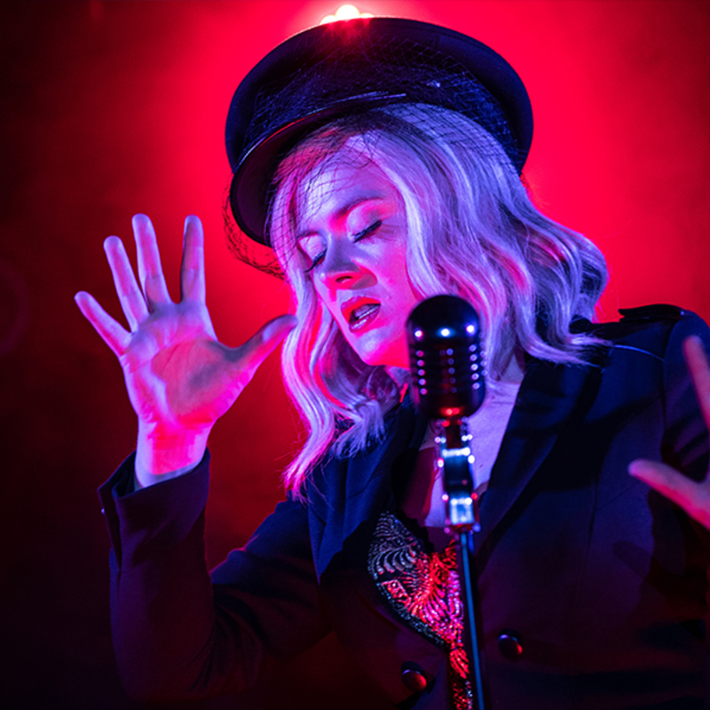 A female performer spreads her fingers wide on either side of a microphone. The female performer has a military style hat, white hair and red lips. Behind her is a red spotlight that frames her stance. She has her eyes closed and is singing.