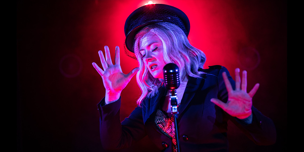 A female performer spreads her fingers wide on either side of a microphone. The female performer has a military style hat, white hair and red lips. Behind her is a red spotlight that frames her stance. She has her eyes closed and is singing.