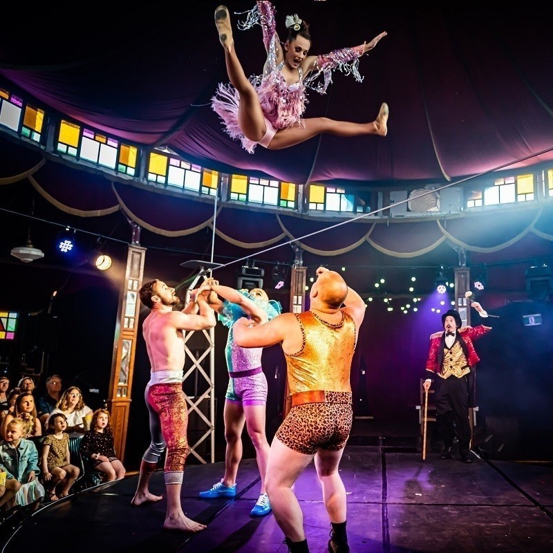 Three acrobats throw a female acrobat into the air. They are colourfully dressed inside the Wonderland Spiegeltent.