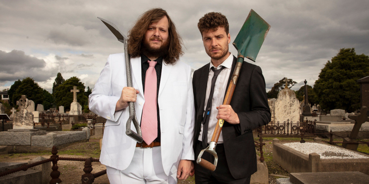 Two men in a grave yard, wearing suits, holding shovels over their shoulders, staring down the barrel of the camera.