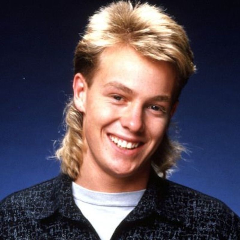 Jon Bennett vs Jason Donovan - A '90s headshot of a young man smiling, he has a blonde mullet and wears a black and grey patterned shirt with a blue background.