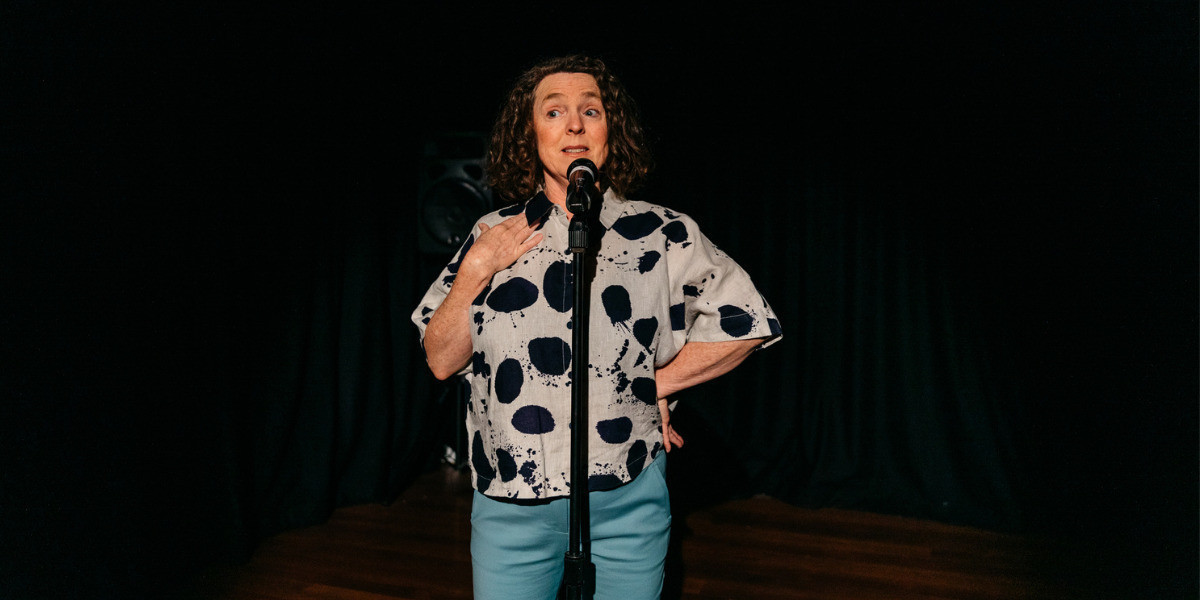 Caroline is on stage and standing behind a mic performing her show. There are no other props on stage with her. She is wearing jeans and a black and white print shirt. She has an amused theatrical look on her face as she speaks.