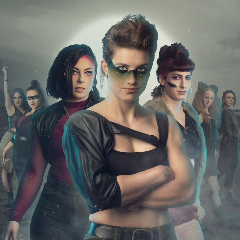 Seven female and gender queer artists stand facing the camera with torn clothes and war paint on their faces with a stark deserted landscape behind them. They have expressions that range from determined to fierce, resigned to playful. The colours in clothes, face paint and background are faded greens, maroon and faded grey.