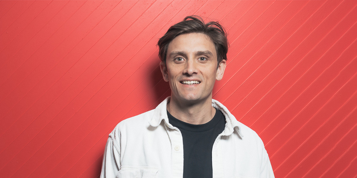 Sam is wearing a black t-shirt and white jacket. He has short brown hair and brown eyes. He is smiling directly into the camera. He is standing in front of a red backdrop that has diagonal lines through it.