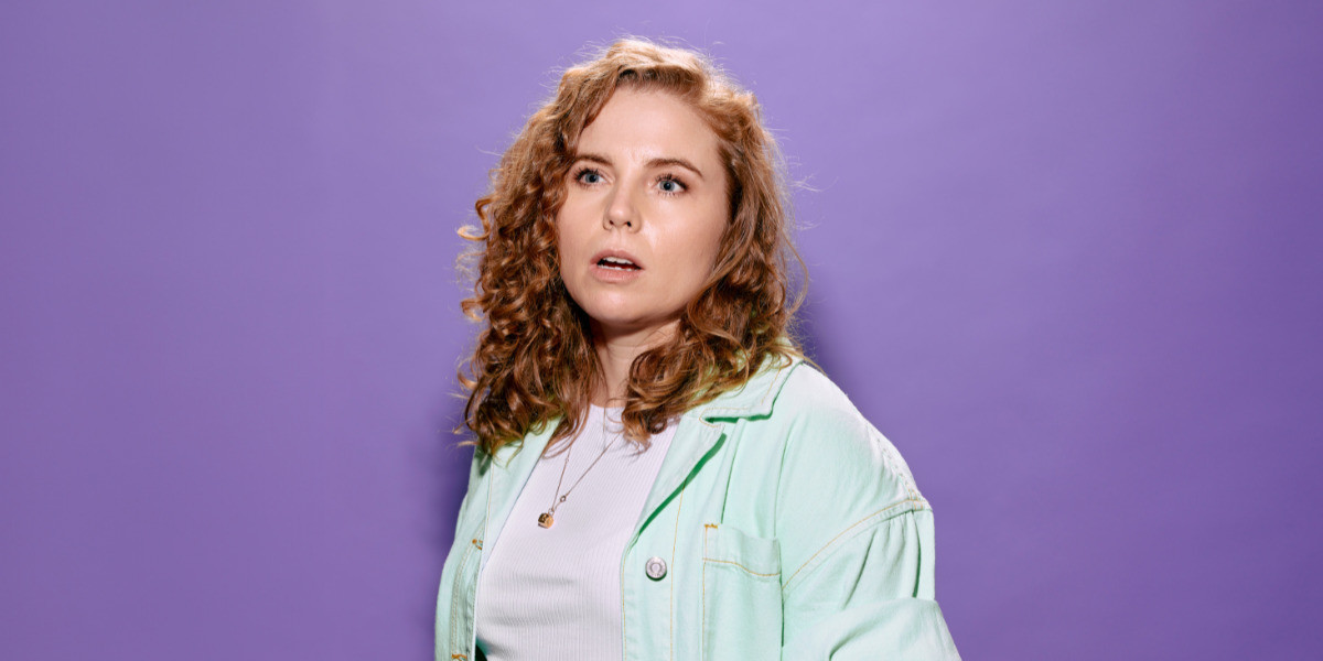 Bronwyn has long strawberry blonde curly hair. She is wearing a white shirt with a mint green button up. Her mouth is gaped and she is looking out of camera. The background is light purple.