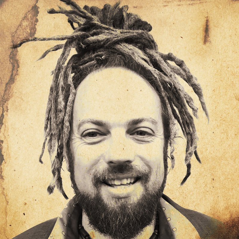 A sepia toned head shot of a man with fair skin and long dreadlocks tied up in a bun on top of his head. He has a short dark beard and a big smile. The background is like in the style of an old fashioned wanted poster.