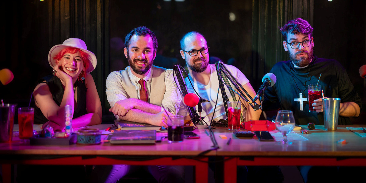 Role To Cast LIVE - The four cast members of Role To Cast pose and smile at the camera from behind their game table at The Lost Dice Cafe. From left to right they are Ellen, Sean, Phil and Chris. They are lit in bright colours and wearing 1930s inspired clothing.