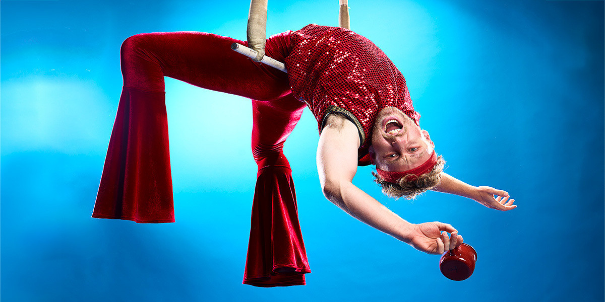 a circus performer dressed in red, dangles from a trapeze. they are against a blue background