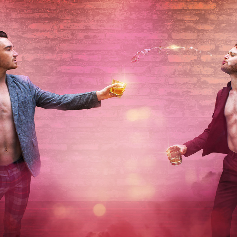 2 people in suit jackets and no shirt hold drinks toward each other on a brick background. One is spitting his out.