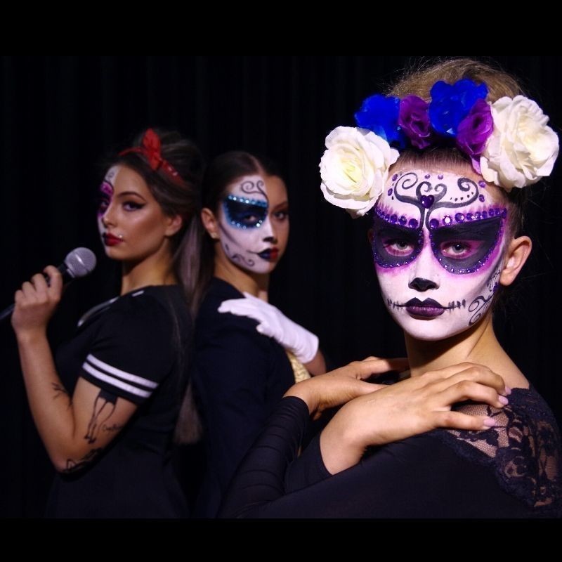 Image of a girl with her face painted as a sugar skull wearing a flower crown.
In the background are 2 girls in costume dressed as Amy Winehouse and Michael Jackson.