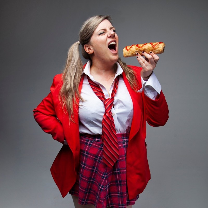 a blonde woman in a red jacket, white shirt and stripy red tie is poised to bite into a sausage roll aggressively