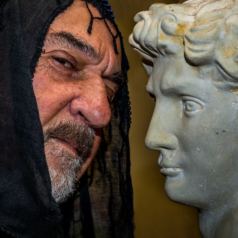 An close up image of a man’s face wearing a black hood next to a stone statue of a person's face.