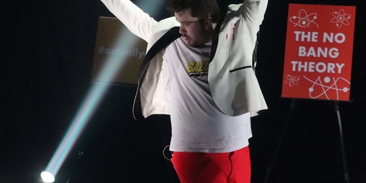 A white man with dark hair and his arms in the air is wearing a white t'shirt with Bazinga on the front and a black trimmed off-white jacket and red trousers. He is looking across his body and down behind him. He is in front of an orange sign that reads The No Bang Theory and there is another sign a bit further back that reads #actuallyautistic - there is a single spotlight beam shining from the ground behind him to the roof.