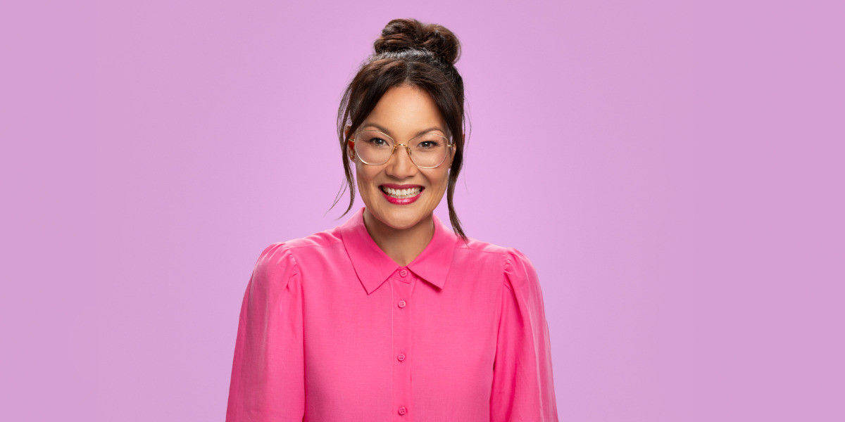 Lizzy has dark brown hair in a top bun. She is wearing big round glasses and a pink button up shirt. Lizzy is smiling directly into the camera. She is in front of a purple backdrop.