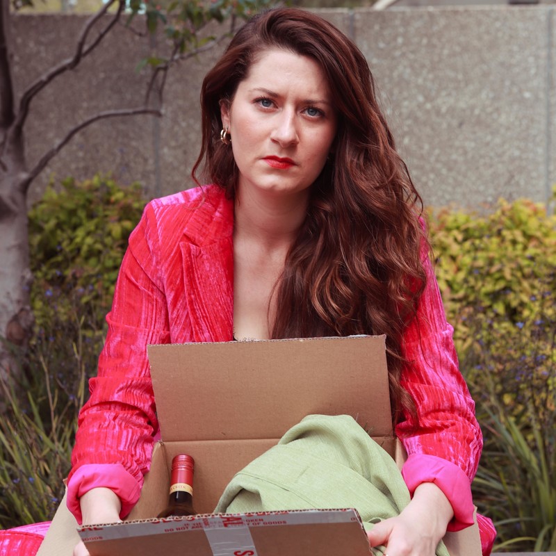 Image of young woman wearing a pink velvet suit, holding a box containing a green jacket and wine bottle. She appears upset and angry.