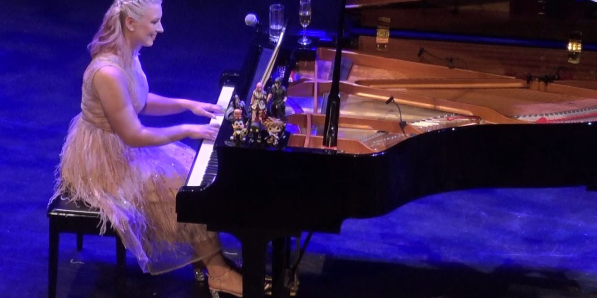 blonde woman with wavy hair smiles while playing a concert grand piano. she is wearing a pink feather dress and there are video game figurines on top of the piano