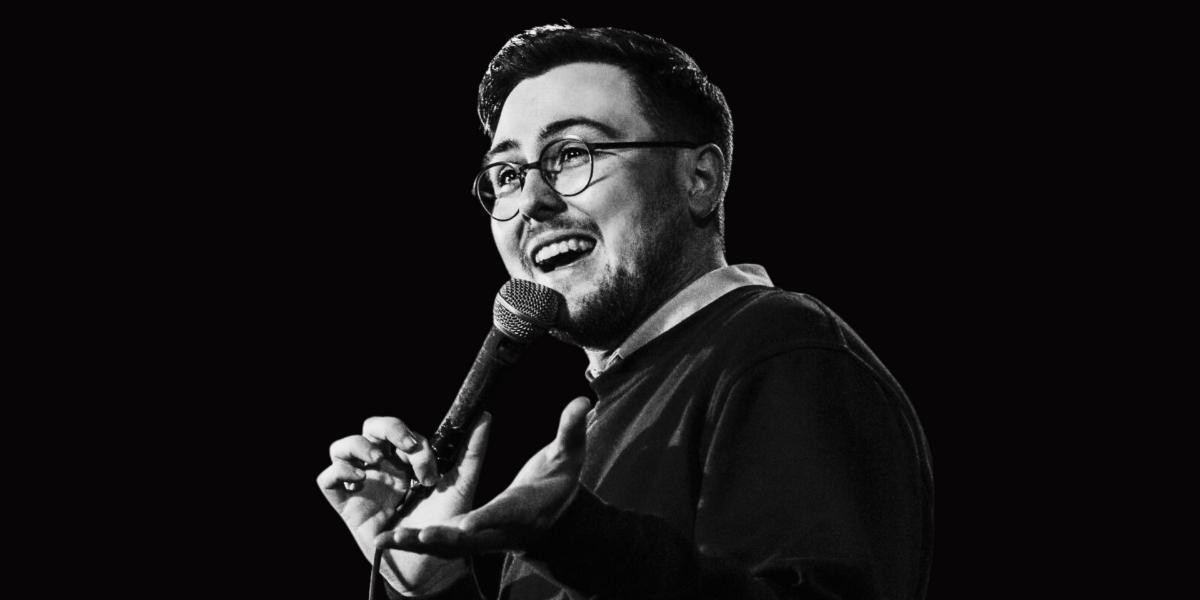 Peter James | Me, Again. Hello. - A black and white image of comedian Peter James on stage, holding a microphone and laughing, looking off camera, and cropped from the shoulders up.