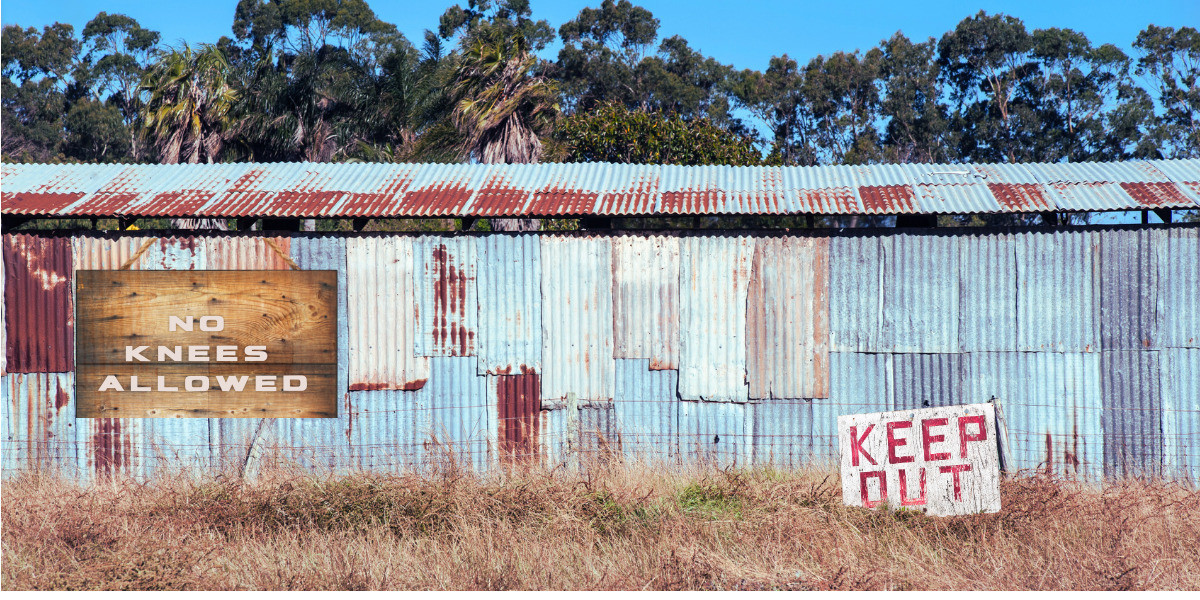 A big shed in Mudbrick - a large tin shed with rusted panels of tin. And a keep out sign in the grass near the shed. A sign that says "No knees allowed"