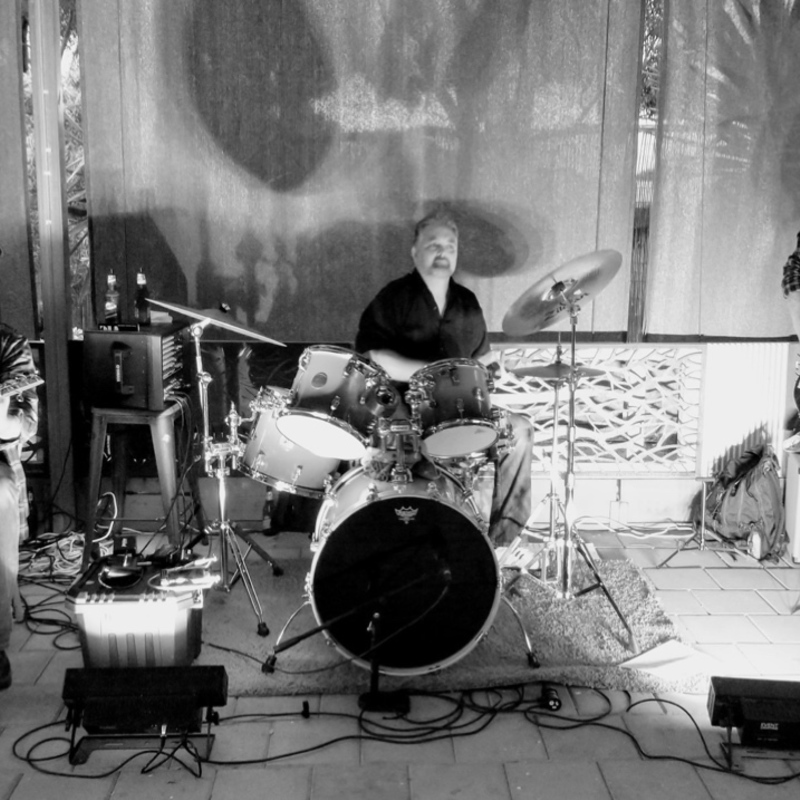 The Brett Littlefair and Devils Bend band are shown consisting of a seated guitarist playing a cigar box guitar, a drummer and bass guitar player standing, in black and white with front upward lighting inferring a bluesy swamp mood related to their music style