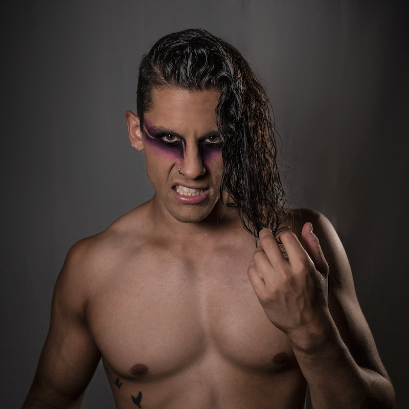 Rouge Goes Rogue - A shirtless man with pink and black theatrical make up looks at the camera with a snarl on his face while twisting a lock of his shoulder length black hair between his fingers.