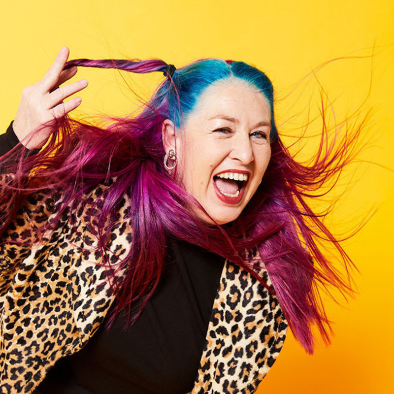 Kelly Mac leans forward against a yellow background. She is Caucasian and has long pink and blue hair blowing around her face. She wears a leopard-print coat and looks off-camera with a wide-mouthed grin.