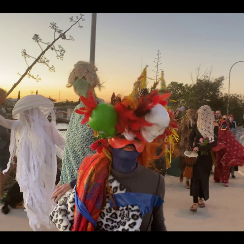 A procession of strange masked figures is crossing the Birkenhead Bridge. The sun is setting in the background.