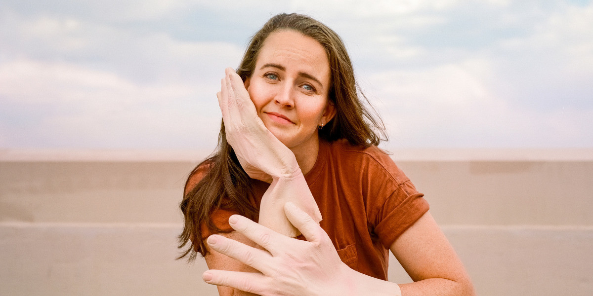 Zoë has long brown hair and blue eyes. She is wearing a burnt orange t-shirt and large hand gloves. The background is a blue sky and the foreground is a dark wooden table. She is smiling at the camera.