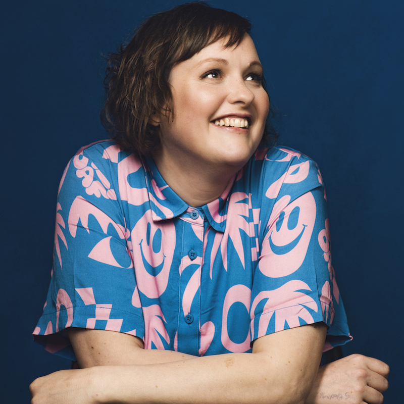Josie is smiling looking to the right of the image wearing a blue top with pink flowers on it. Her arms are folded in front of her as she leans on a desk, shoulders slightly up. She has short brown hair.