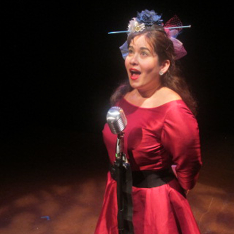 Feisty brunette in a red dress, wearing a knitting hat and singing in a microphone.