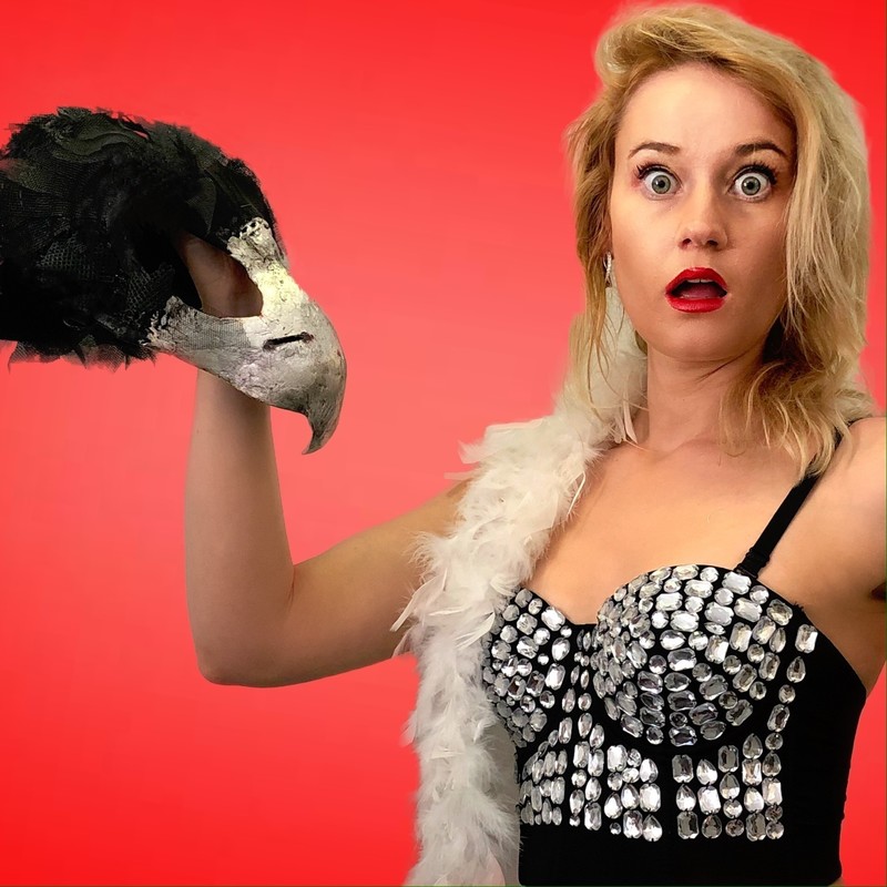 Bird Be Crazy - A photo of a woman with a shocked expression on her face holding a costume mask of a bird’s head. The woman has blonde hair that sits above her shoulders and is wearing red lipstick. She has a white feathered scarf over her shoulder and is wearing a structured black corset singlet with silver embellishments.