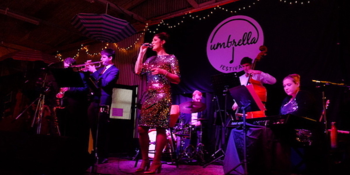 Band on stage at Umbrella festival, female lead singer in sequinned dress, key board player  female in sequinned dress, men playing their instruments drums, saxophone, double bass and trumpet, dark mood lighting.