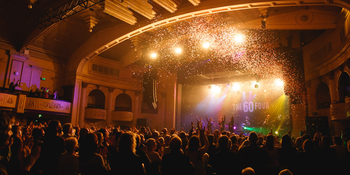 A lively concert scene inside a classic theater. The band name 'The 60 Four' is lit up on stage with performers actively engaging the crowd. Confetti showers down, adding to the festive atmosphere. The audience is fully immersed, clapping and raising their arms in response. Spectators fill the balconies, showcasing a packed house.