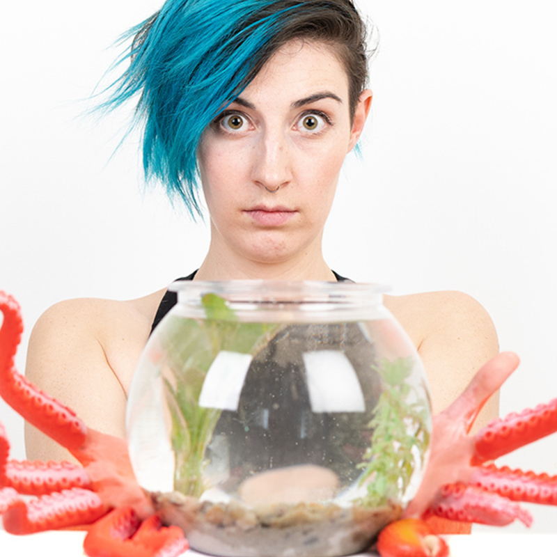 A person with blue pixie-cut hair has pink-orange tenticles instead of hands. They are holding a small fish bowl in front of them. White background.