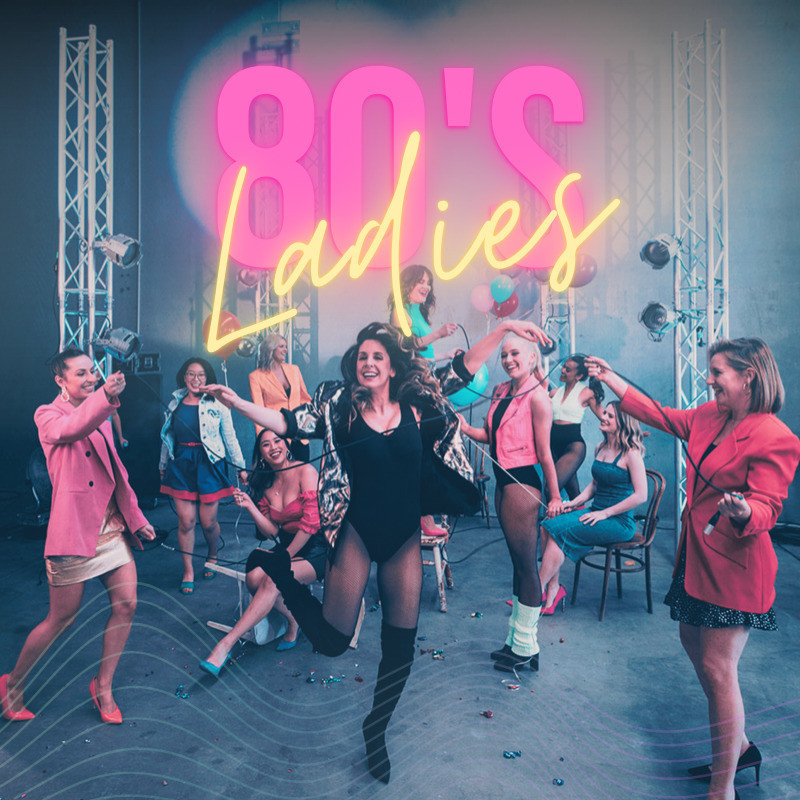 Image of a group of women, wearing 80's inspired costumes, are candidly dancing, laughing, skipping rope (with instrument leads) and jumping around. The title of the show "80's Ladies" is in bold neon pink and yellow is up top.