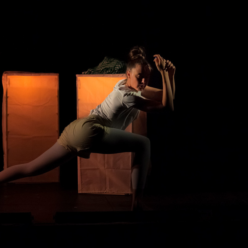 The performer is in a long, low lunge, with her arms held gracefully crossed in front of her. She appears on a black stage with glowing golden set pieces behind her. They are rectangular cube drawers with a light on the inside. She wears yellow shorts and a white t-shirt.