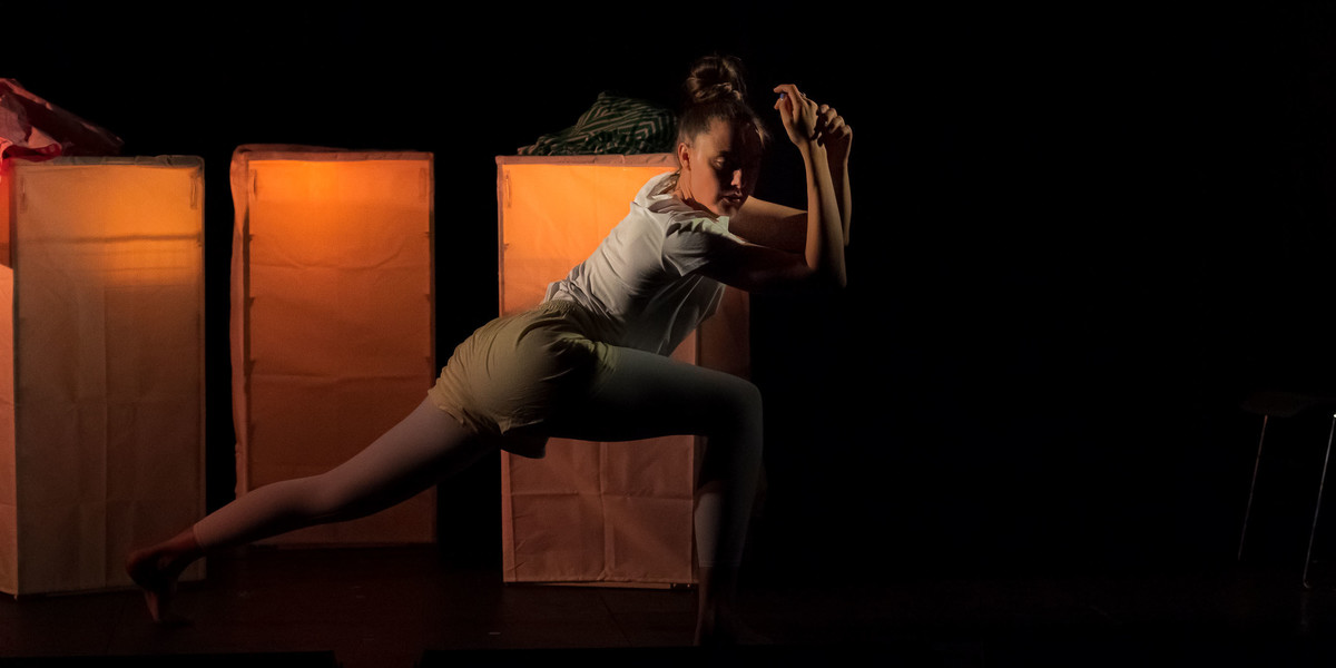 The performer is in a long, low lunge, with her arms held gracefully crossed in front of her. She appears on a black stage with glowing golden set pieces behind her. They are rectangular cube drawers with a light on the inside. She wears yellow shorts and a white t-shirt.