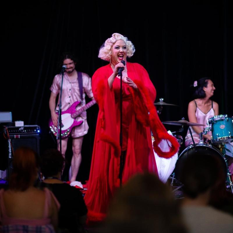 Performance of Endo Days, with Libby Trainor Parker centre stage and smiling wearing a red dress and blonde wig. Behind her is the Jarmy Army band in pink pyjamas.