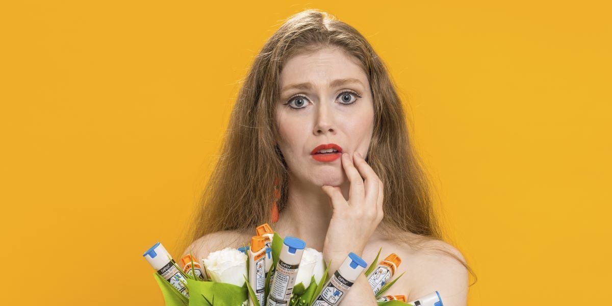 Intolerant - Uma is pictured from the elbows up, holding a bouquet of epipens and flowers, and using her right hand to touch her face, looking concerned. The background of the picture is a golden yellow, and Uma is wearing her strawberry-blonde hair out, with a blue formal dress.