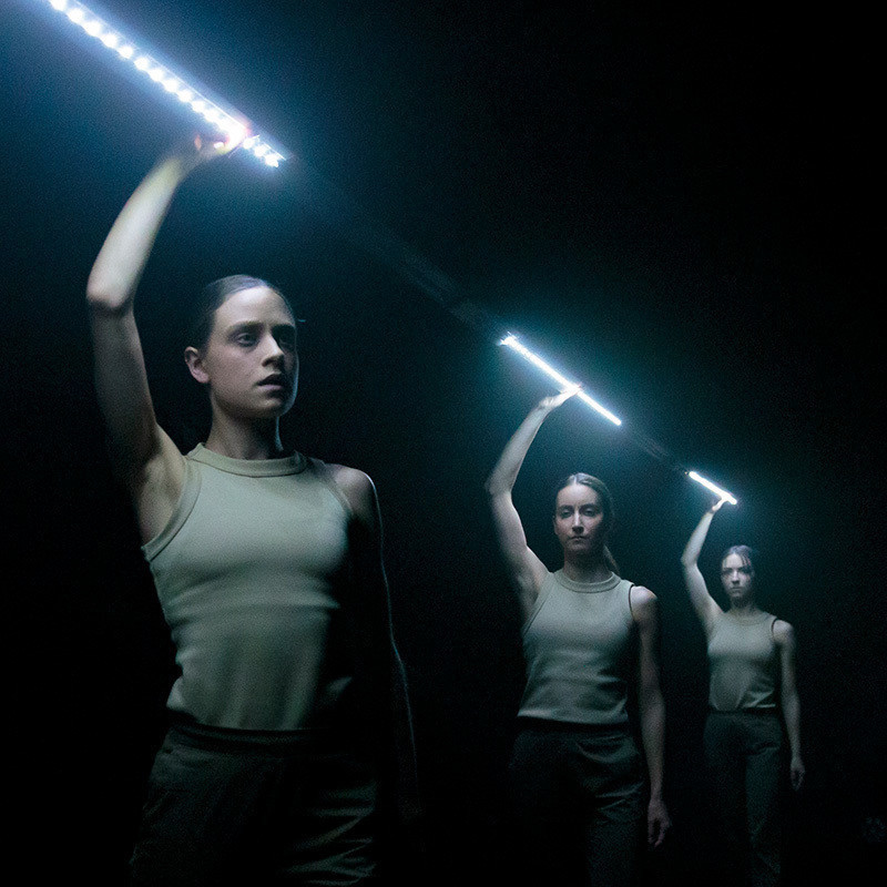 Three performers wearing khaki singlets and pants standing in a line on a darkened stage. Their faces are illuminated by small white lights attached to a long bar which they hold overhead with one arm.