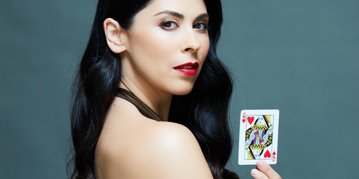The female magician holds a playing card in her hand. She has long dark brown hair and a green dress with red lipstick on her lips.