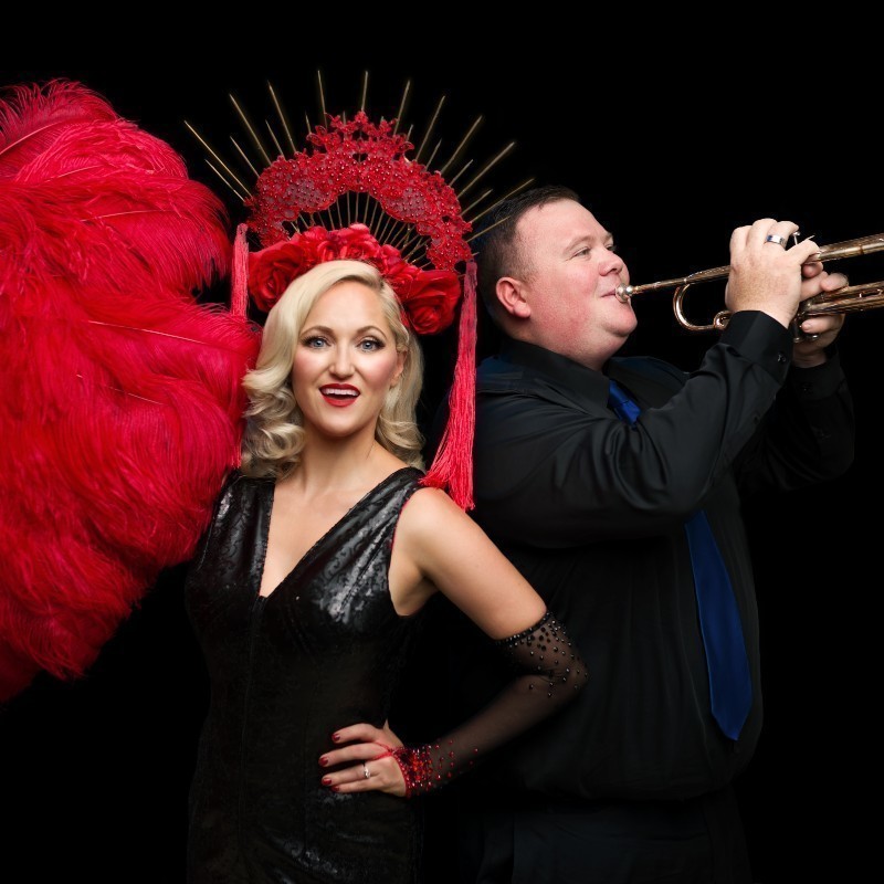 Swingin' Burlesque - A photo of a woman with blonde hair wearing a black dress and extravagant red headpiece holding a large red feathered hand fan. The man next to her is playing trumpet and is wearing a black shirt and blue tie.